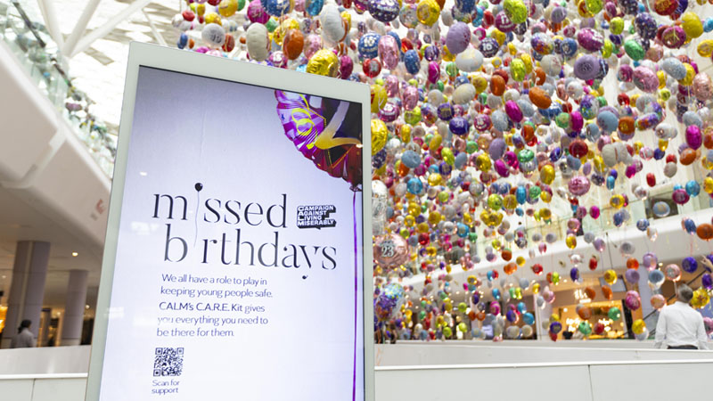 A screen showing the Missed Birthday installation logo and details, stands in front of thousands of colourful, floating balloons