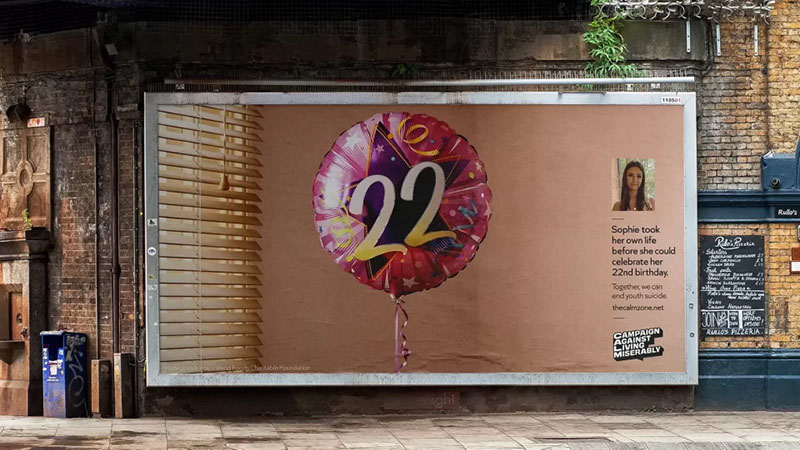 The image shows a billboard by Campaign Against Living Miserably (CALM). Featuring a large balloon featuring the number "22." To the right, there's a photo of a young woman and a message that reads: "Sophie took her own life before she could celebrate her 22nd birthday. Together, we can end youth suicide."