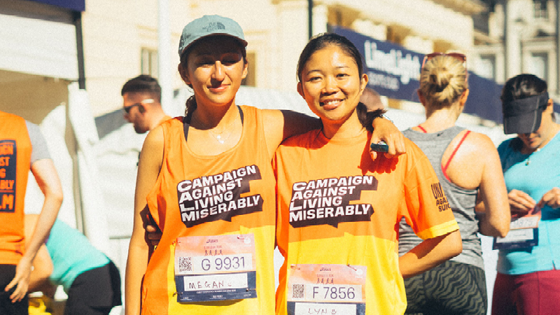 Two women stand together smiling, wearing bright orange and yellow running vests with the "Campaign Against Living Miserably" logo printed on them. 