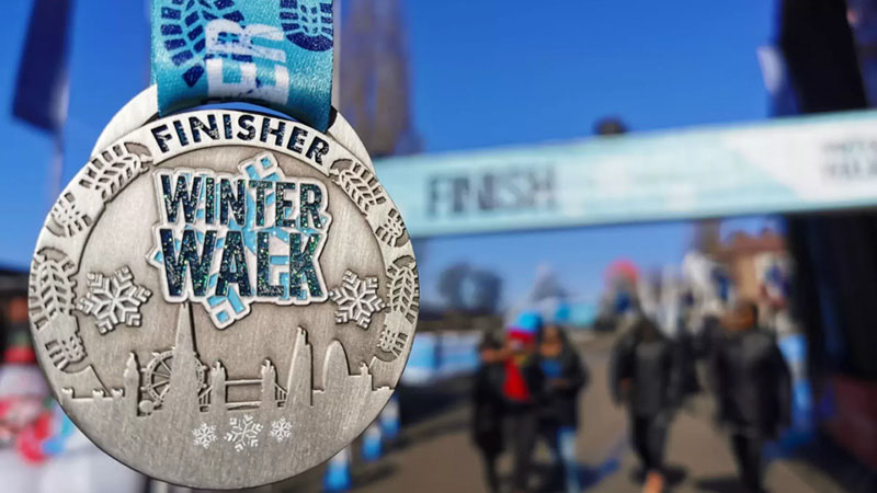 A close-up of a silver medal with the text "FINISHER" and "WINTER WALK" engraved, featuring snowflakes and a city skyline design. The medal is attached to a blue and white ribbon with winter-themed patterns. In the blurred background, participants in winter attire are seen walking near a finish line banner under a clear blue sky.