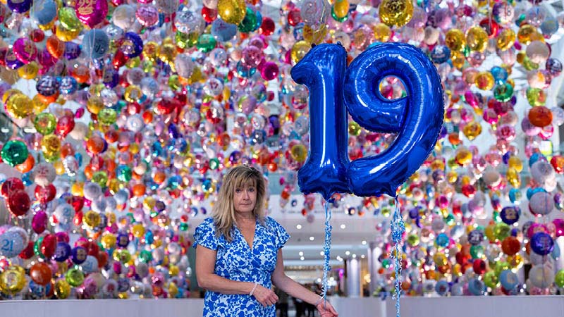 A participant at the Missed Birthdays exhibition, holding large blue balloons shaped like the number "19." Behind her is a colourful backdrop filled with hundreds of suspended, multi-coloured balloons.