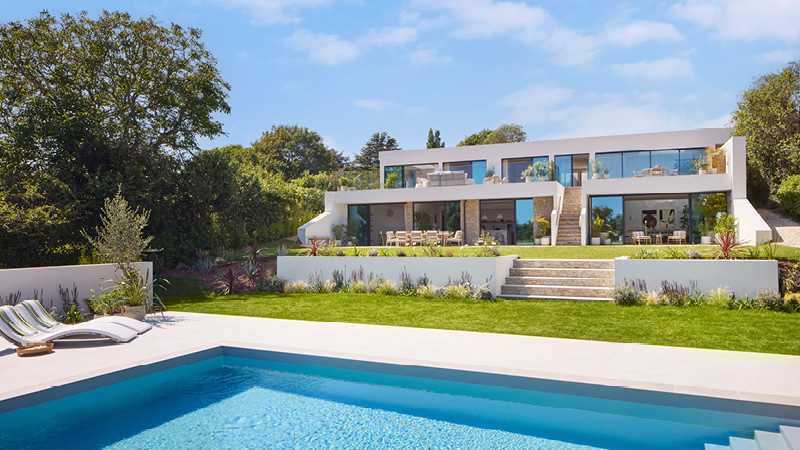 The CALM Omaze house is modern, luxury house with a sleek, minimalist design, featuring large glass windows and terraces on two levels. The house is surrounded by greenery, including manicured lawns, shrubs, and tall trees in the background. A pristine swimming pool in the foreground reflects the bright blue sky, with lounge chairs placed near the poolside for relaxation.